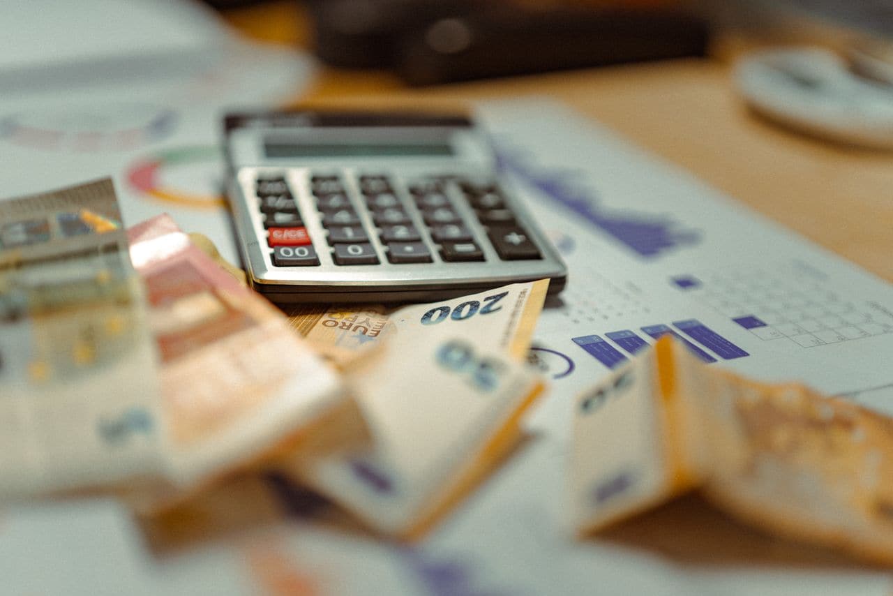 Calculatrice posée sur des tableaux financiers, entourée de billets en euros — le devis d'un projet, avant qu'on ne sache ce qu'il contient.