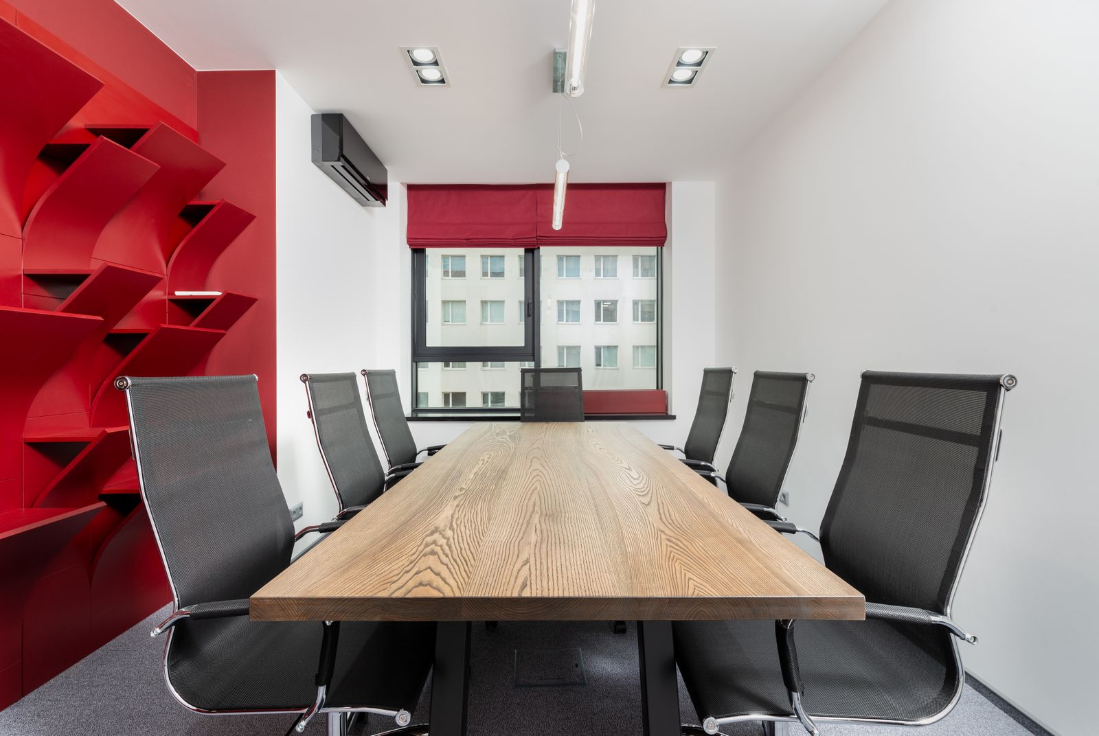 Salle de réunion moderne inoccupée : table en bois, chaises et étagères rouges devant une fenêtre à stores.