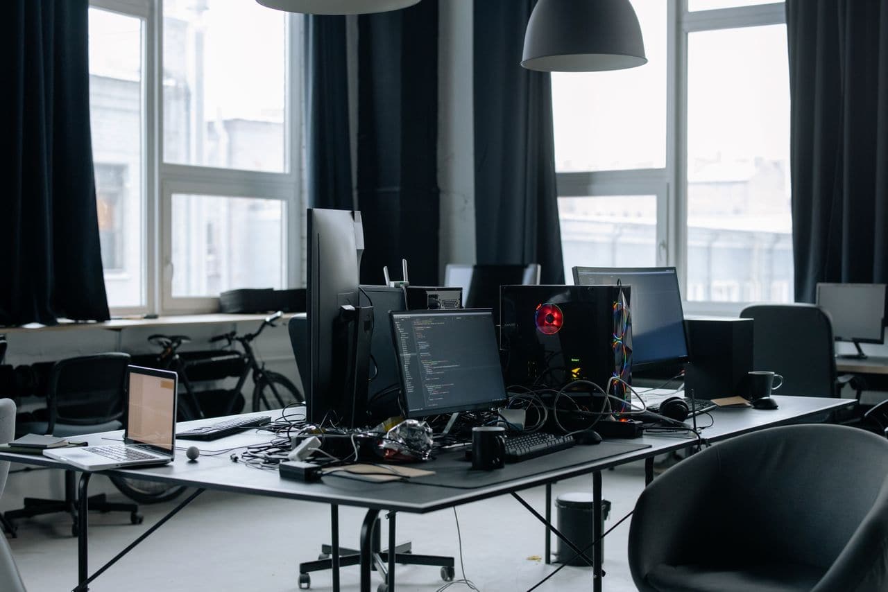 Bureau d'un espace de travail technique désert : écrans allumés affichant du code, chaises vides, poste de direction inoccupé.