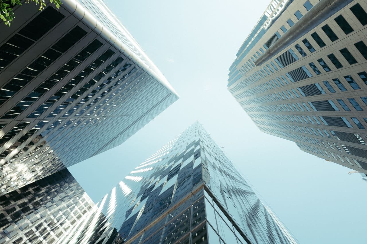 Contre-plongée sur des tours de bureaux vitrées, vues depuis le sol — la posture de celui qui lève les yeux vers une structure qu'il ne maîtrise pas.
