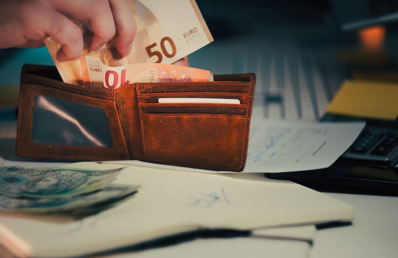 Main sortant des billets d'un portefeuille posé sur des documents de travail — le moment où il faut décider du montant d'une offre, sans repère.