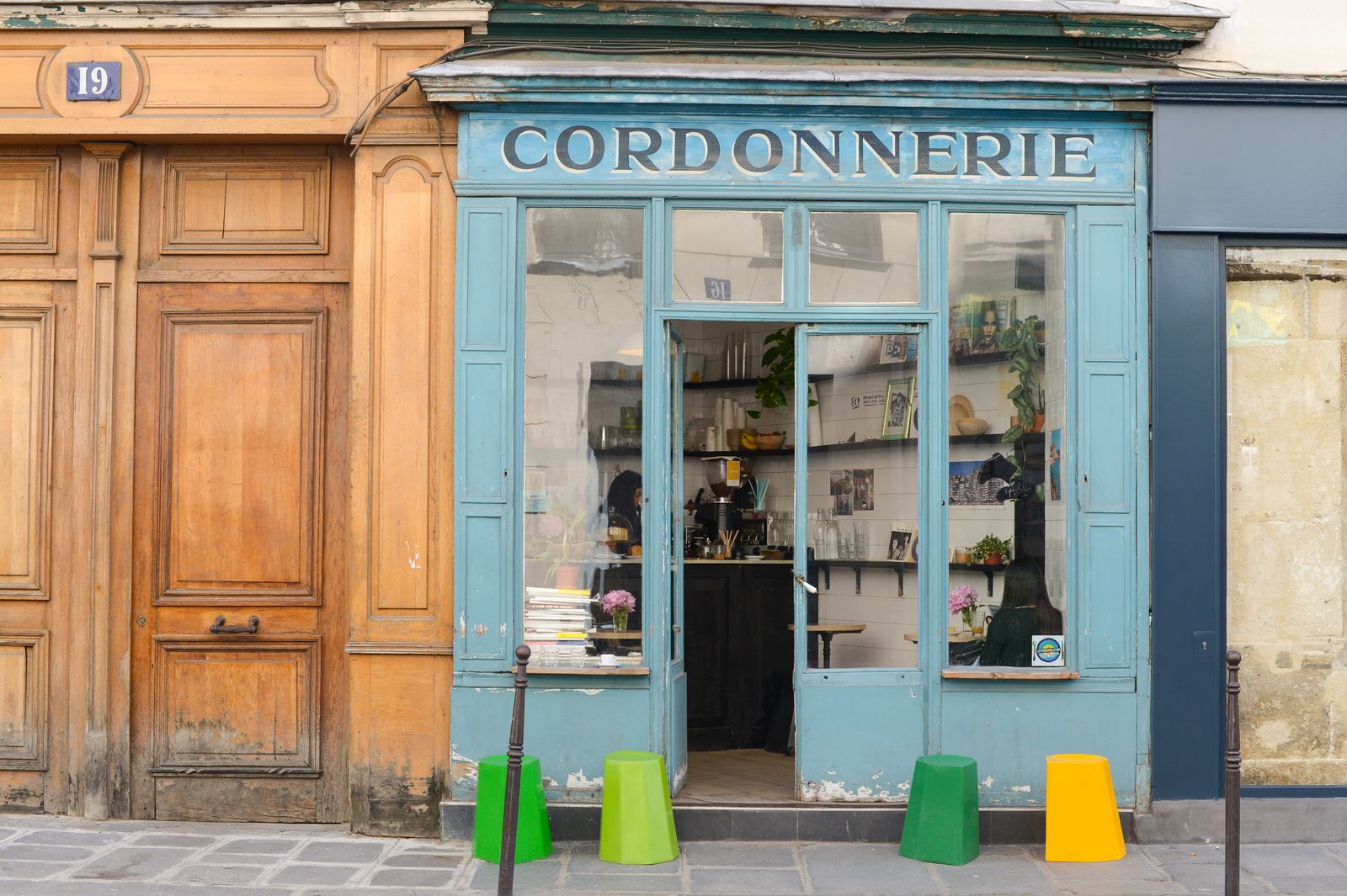 Devanture d'une cordonnerie de quartier, enseigne peinte et tabourets colorés sur le trottoir.