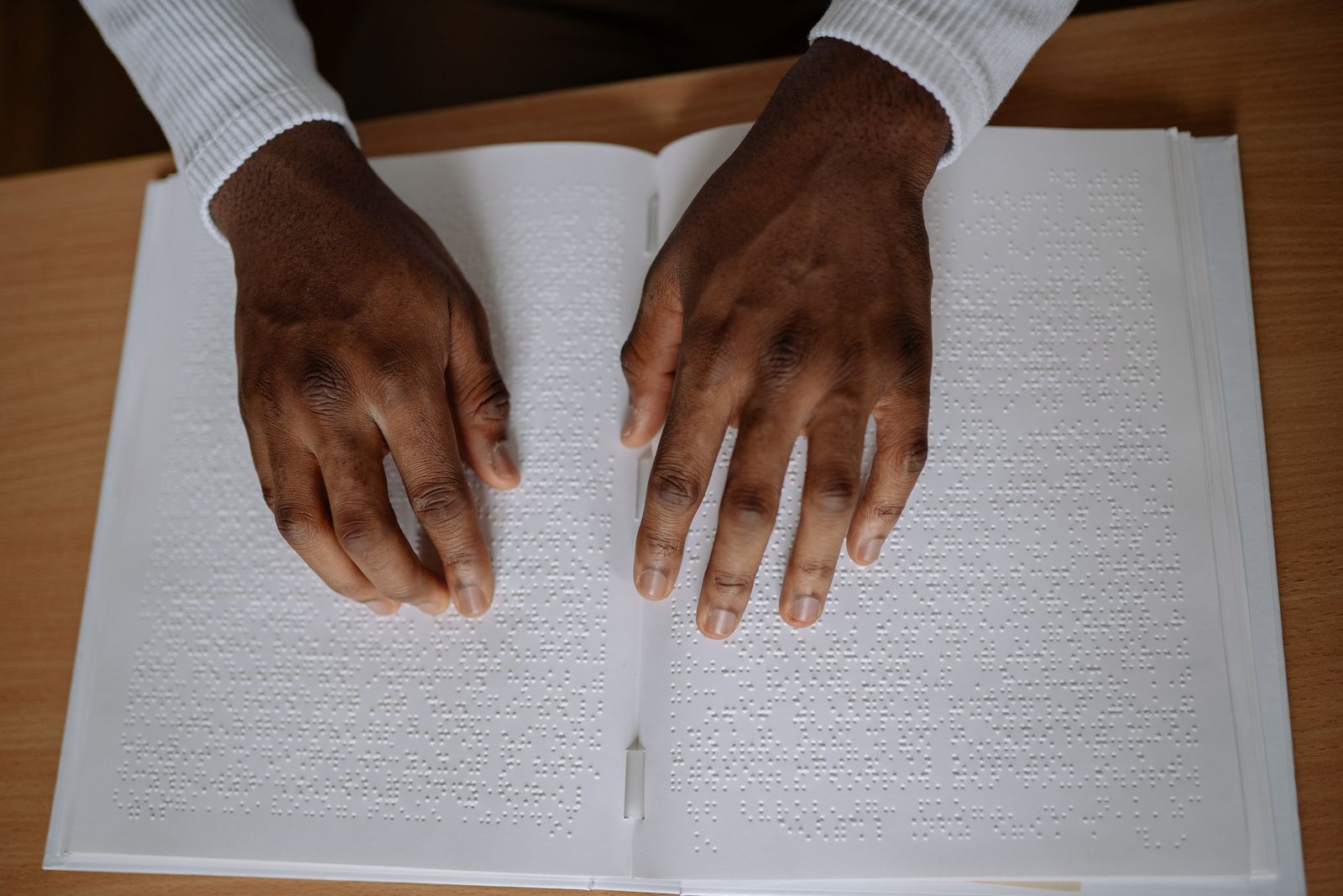 Mains parcourant du bout des doigts une page imprimée en braille, sur une table en bois.