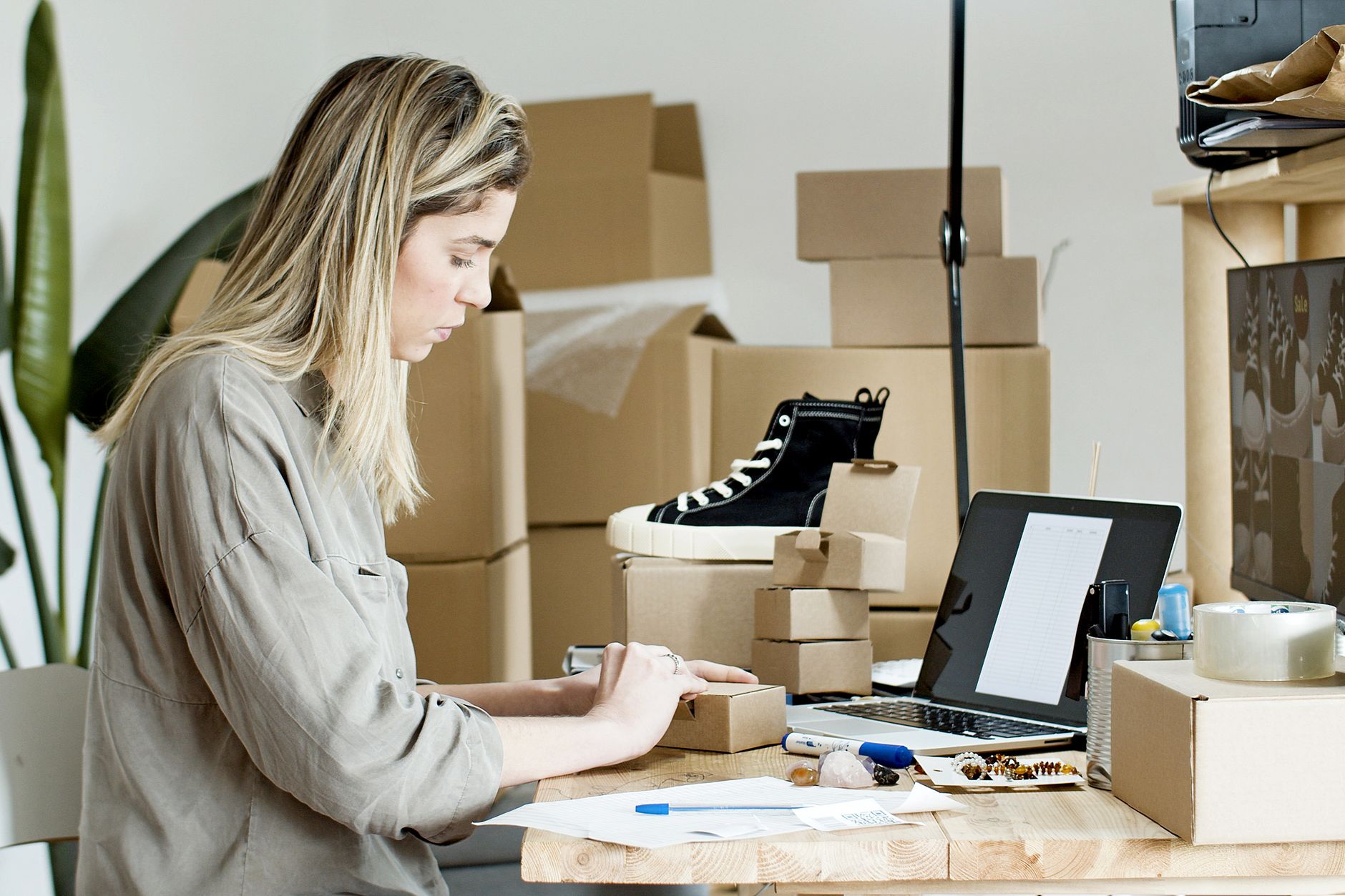 Une commerçante prépare un colis entourée de cartons et de matériel d'emballage, un ordinateur portable ouvert à côté d'elle.